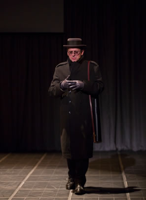 Belikov in black overcoat over black pants and shoes with black gloves and black boater and black scarf hat and a black tube case strapped voer his shoulder walks stiffly